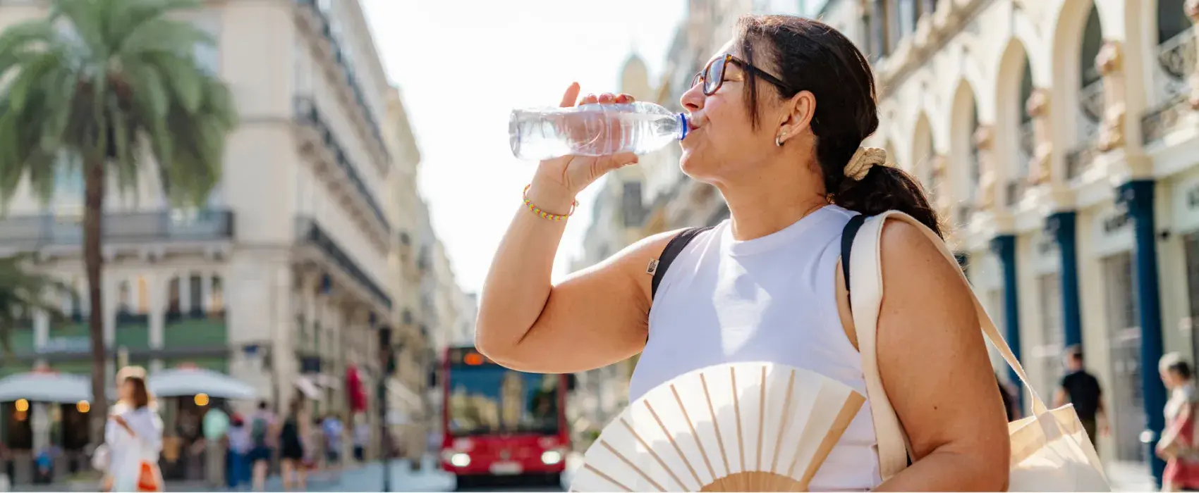 Woman with dark hair and white top drinking from a water bottle and holding a fan in a city