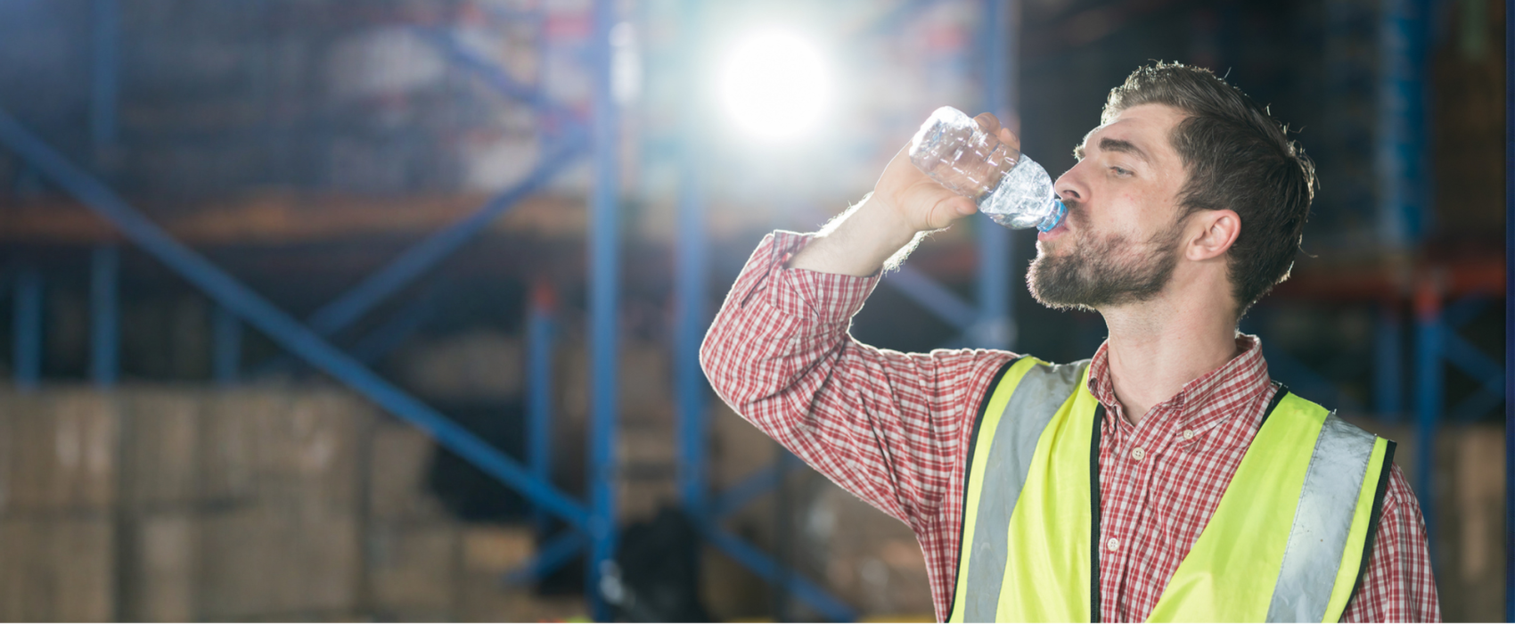 A man with dark brown hair in a red shirt and yellow high vis jacket drinking a bottle of water in a warehouse with the sun shining through
