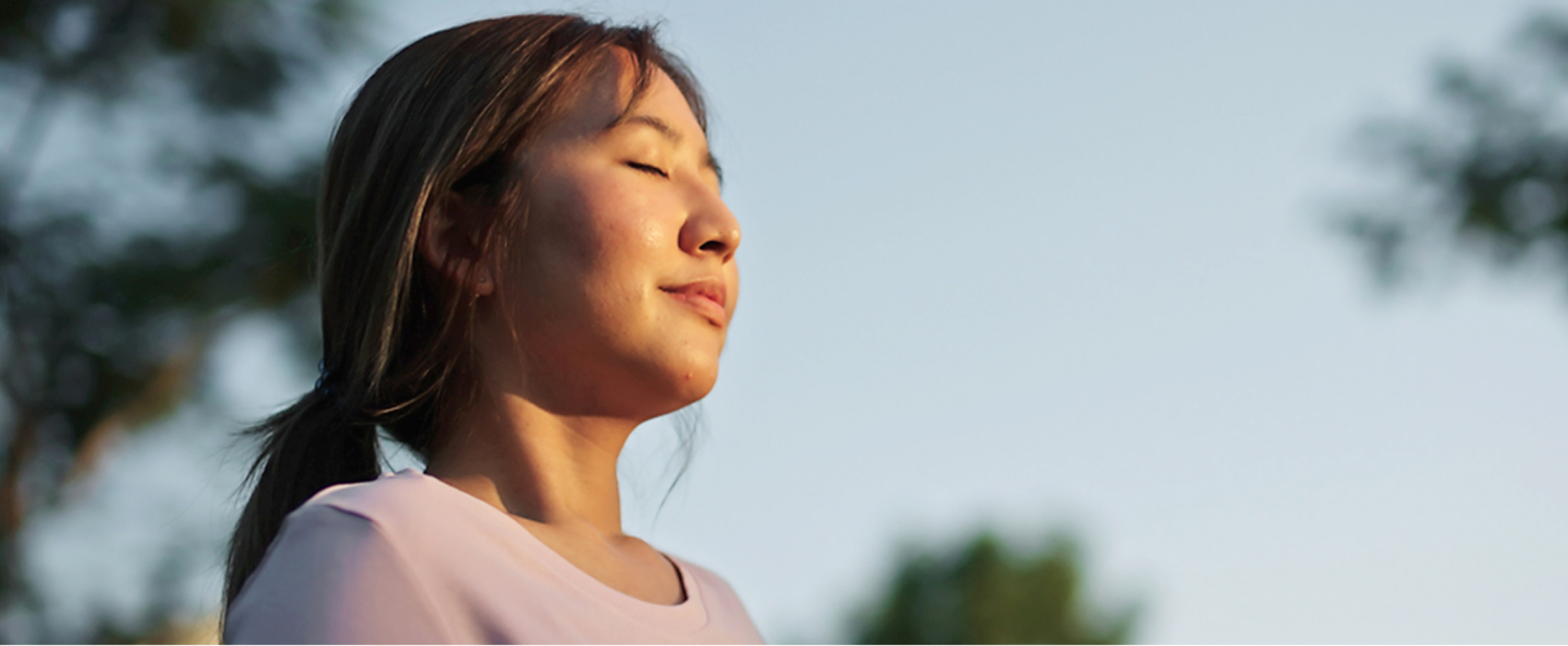 A women wearing a pale pink top with her eyes closed and the sun shining on her face