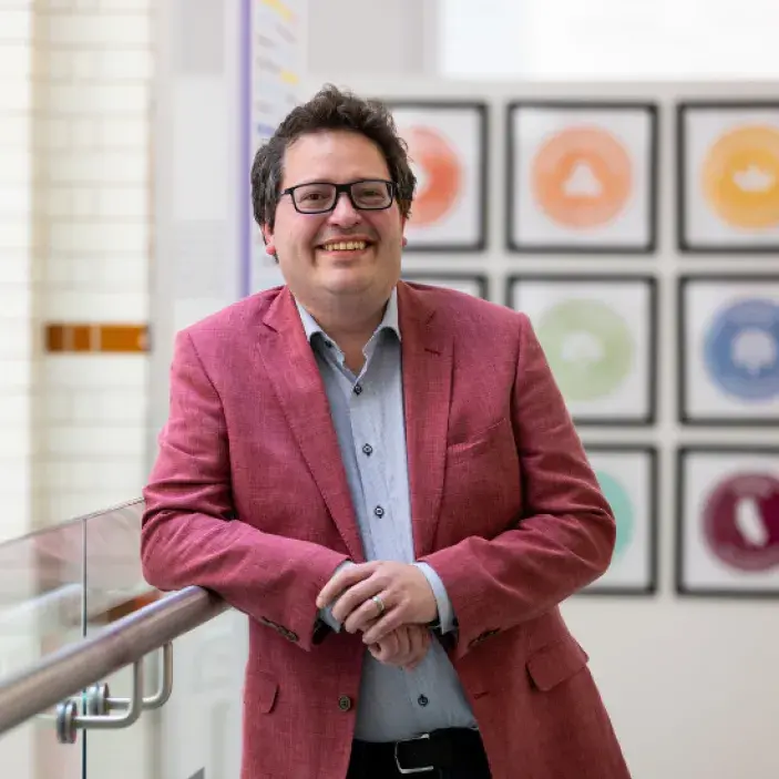 Dark-haired man in a red suit smiling broadly and leaning against a bannister