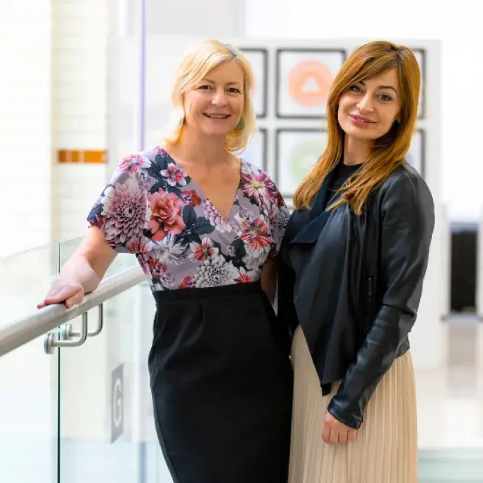 Two women standing in a well-lit corridor looking at the camera and smiling