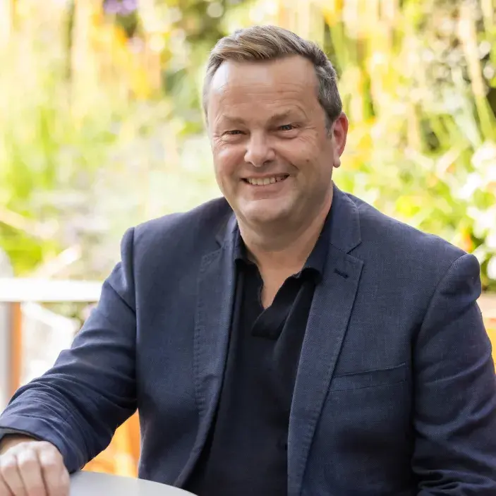 Simon Miles, wearing a dark suit and smiling at the camera whilst sitting at a table outside