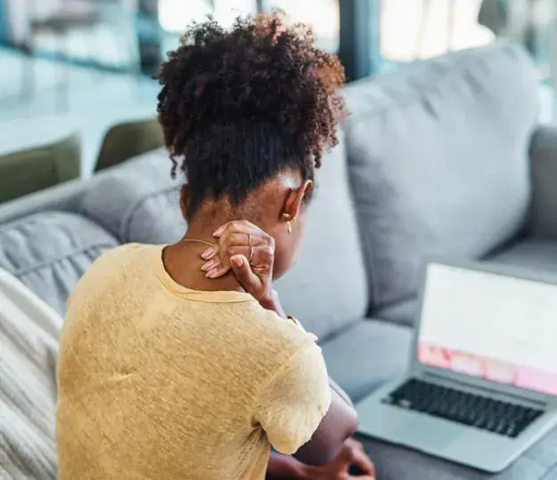 Woman on a couch with a laptop, visibly in discomfort as she rubs her neck