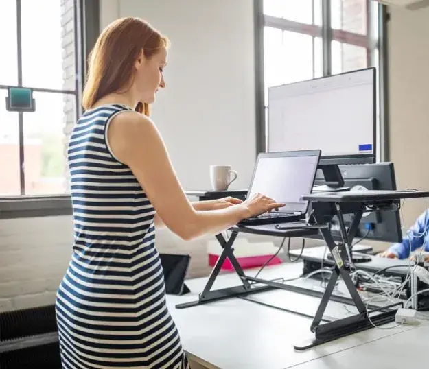 A woman working on her laptop using a standing desk in an office