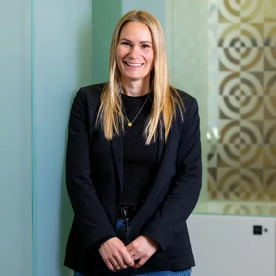 A blonde woman in a black suit standing in a pale blue room, smiling at the camera